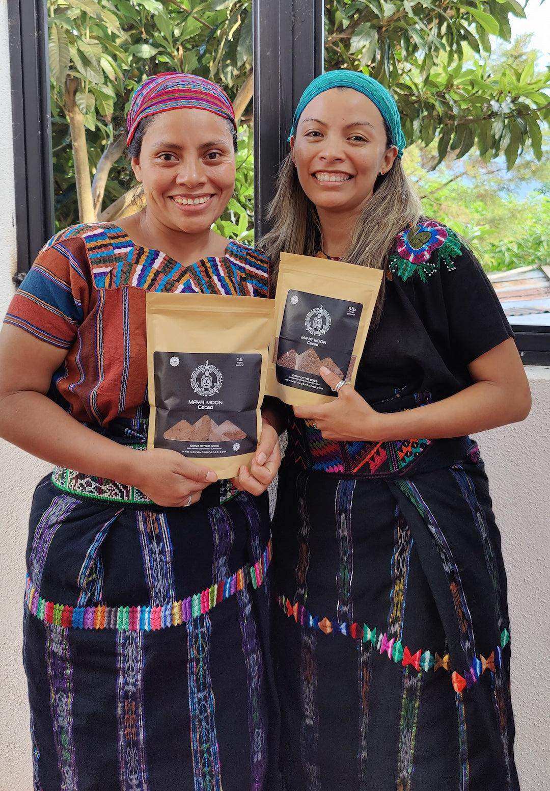 Two women in traditional Mayan attire holding ceremonial cacao bags from Guatemala, smiling outdoors.