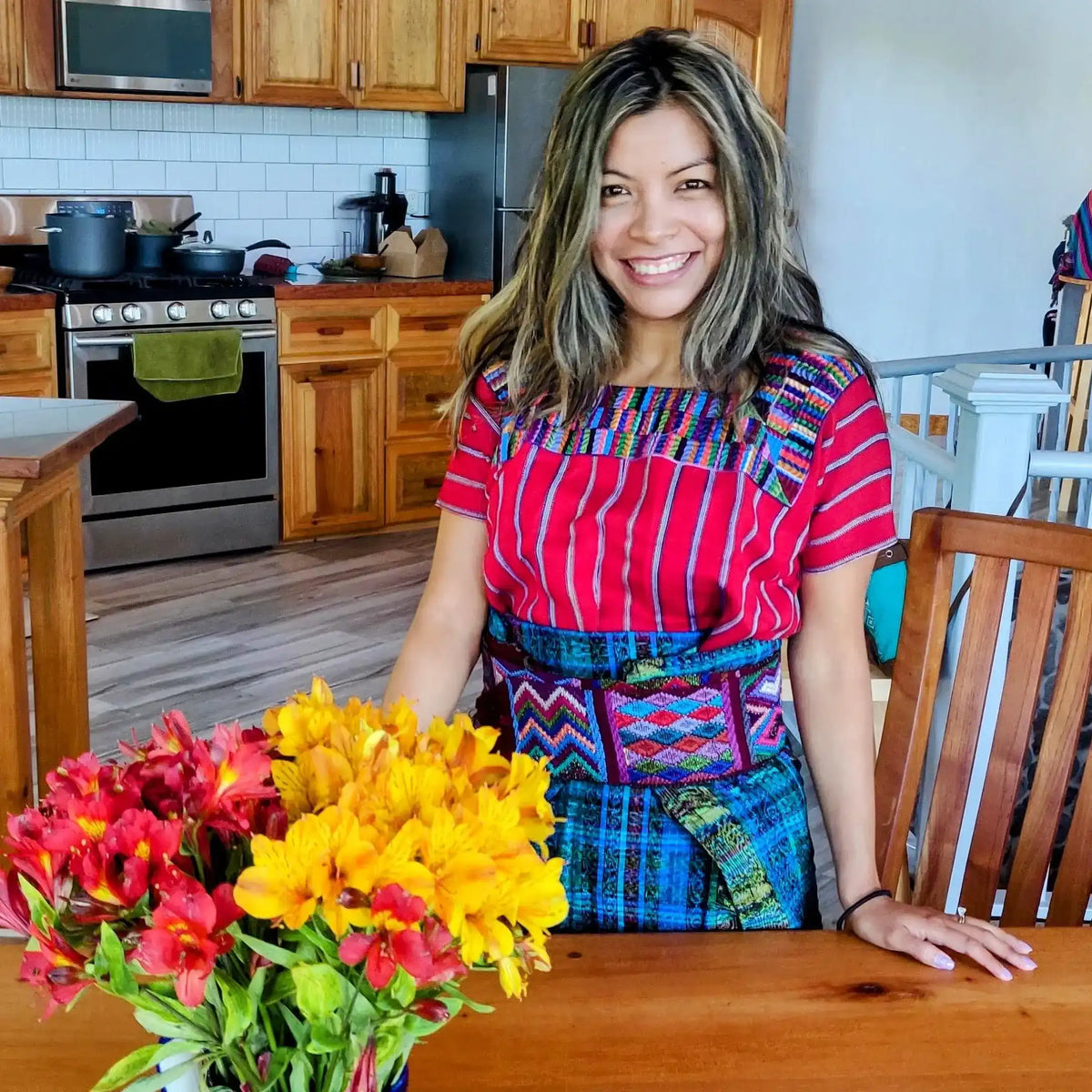 Woman in colorful Mayan attire smiles at kitchen table with flowers, representing Mayan cacao heritage