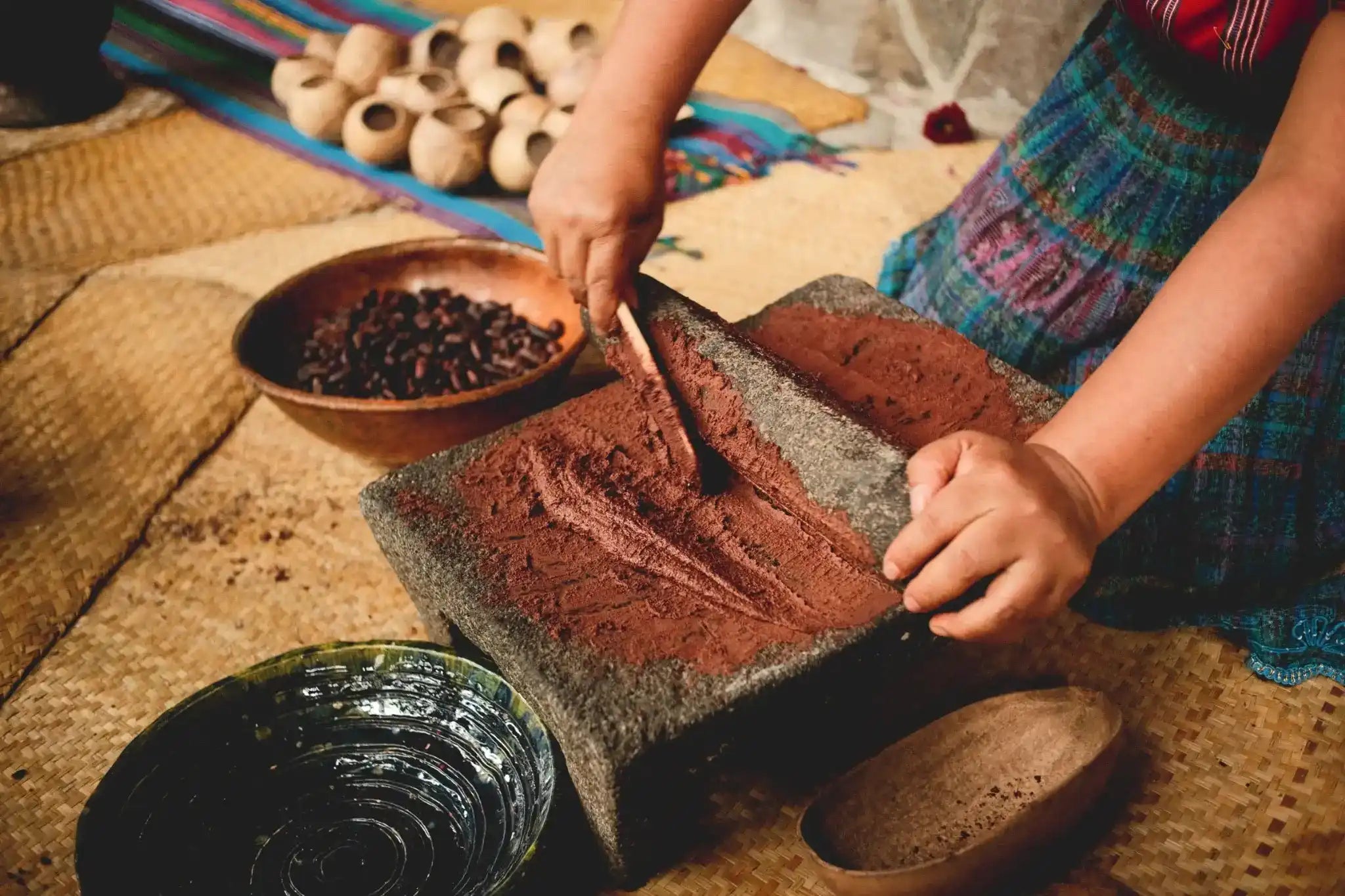 A person's hands using a wooden pestle to grind raw cacao beans into ceremonial pure cacao powder inside a large stone mortar, with bowls of ingredients and dried pods nearby on a woven mat.