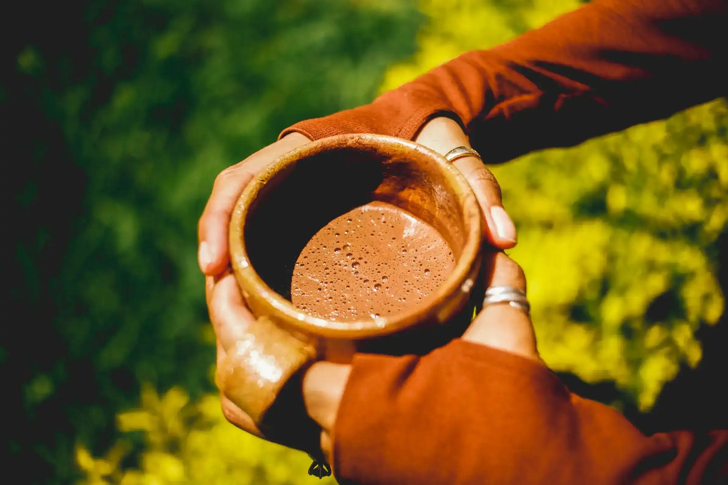 A close-up of a person's hands holding a rustic, handcrafted brown ceramic mug filled with a frothy brown raw cacao powder drink, set against a blurred green outdoor background.
