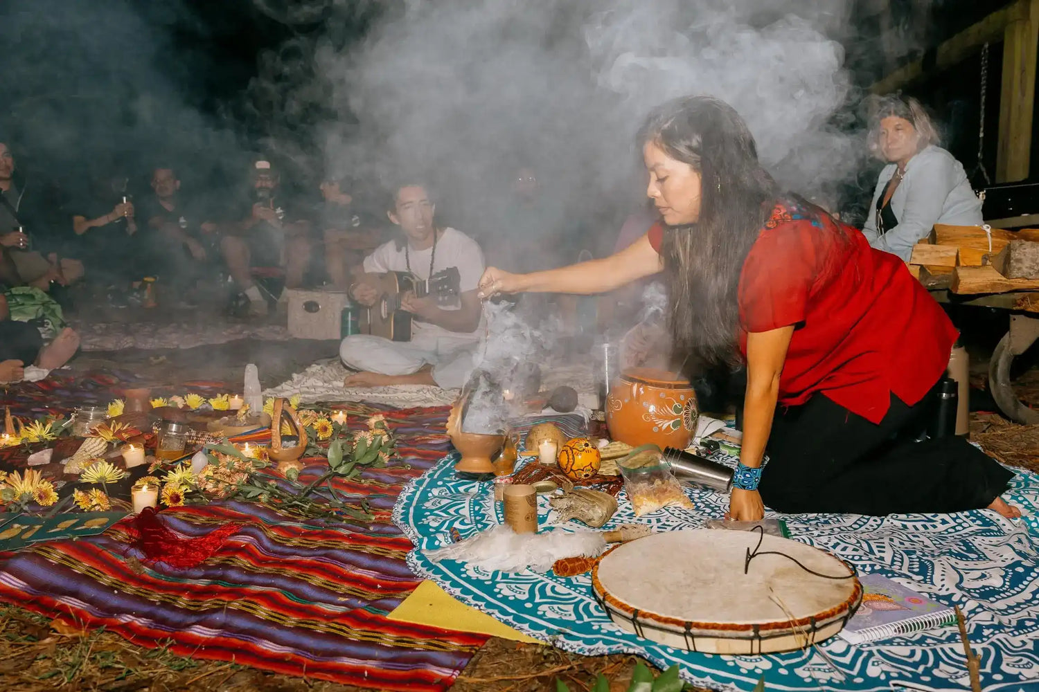 A woman in a traditional Mayan red top and black pants kneels for a smoky Mayan cacao ceremony on patterned textiles, holding a small vessel while others sit in the background in a dim indoor setting.