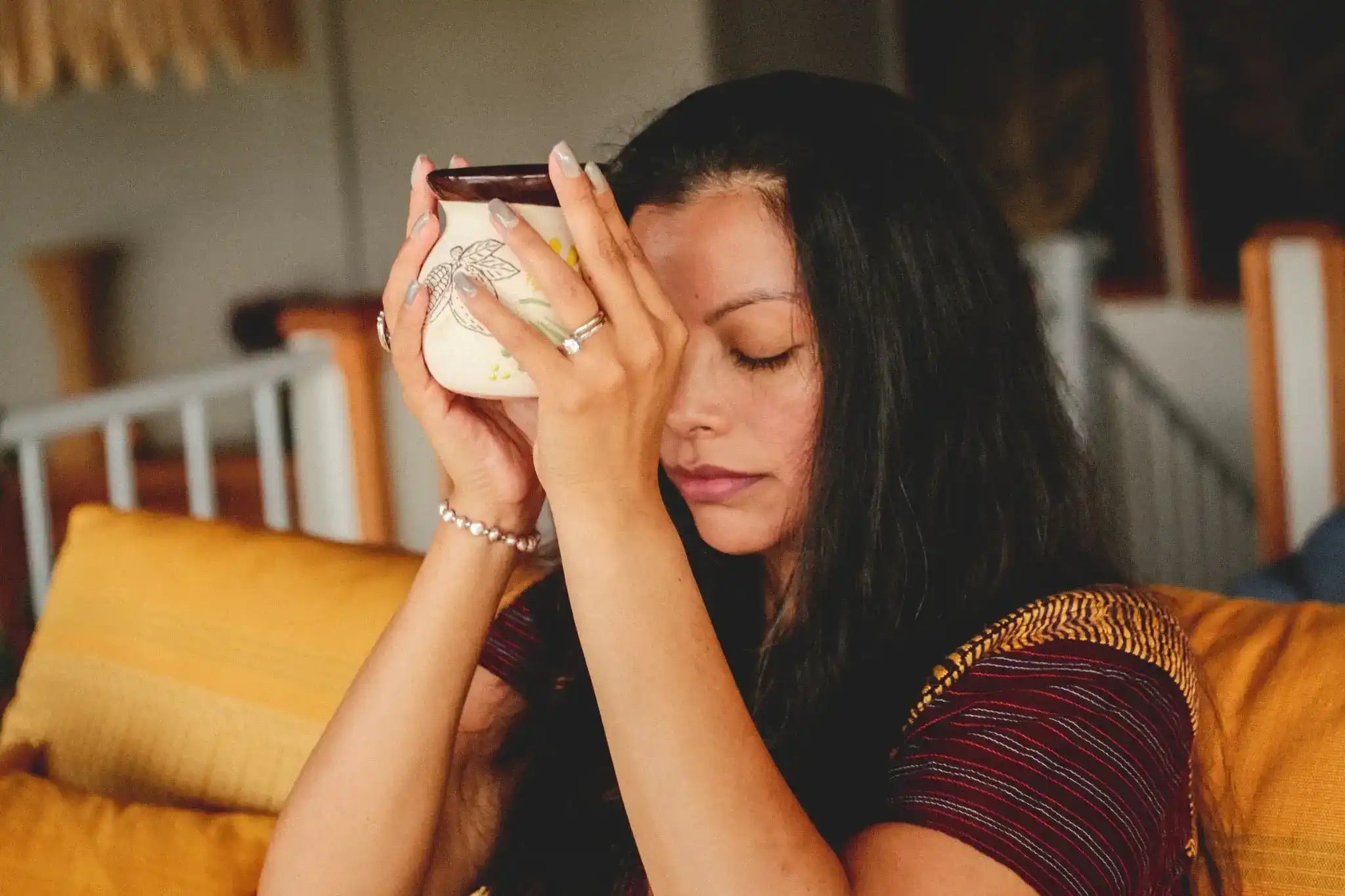 A Mayan woman with long dark hair sits on a yellow couch, holding a small white mug of ceremonial raw cacao powder drink to her forehead with both hands performing an authentic cacao ceremony, while wearing traditional Mayan garb of burgundy top with gold trim and rings on her fingers.