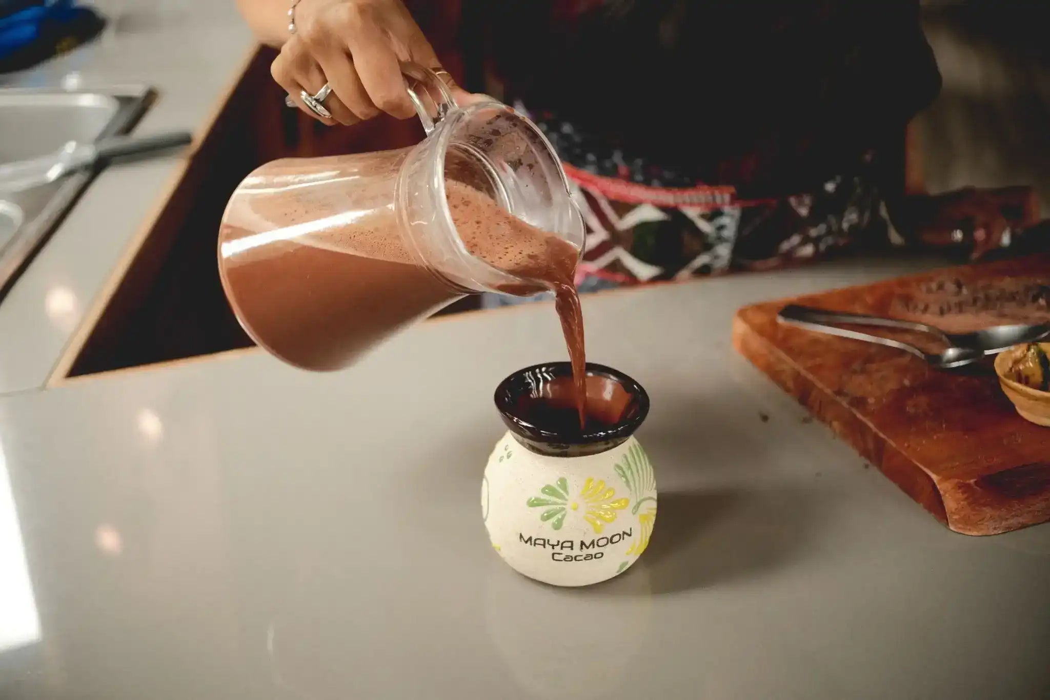 A person pours a dark chocolate alternative pure ceremonial cacao drink from a clear plastic pitcher into a small white ceramic mug with yellow floral illustrations and the text 'MEYA MOON CACAO' placed on a light gray countertop in a kitchen setting.