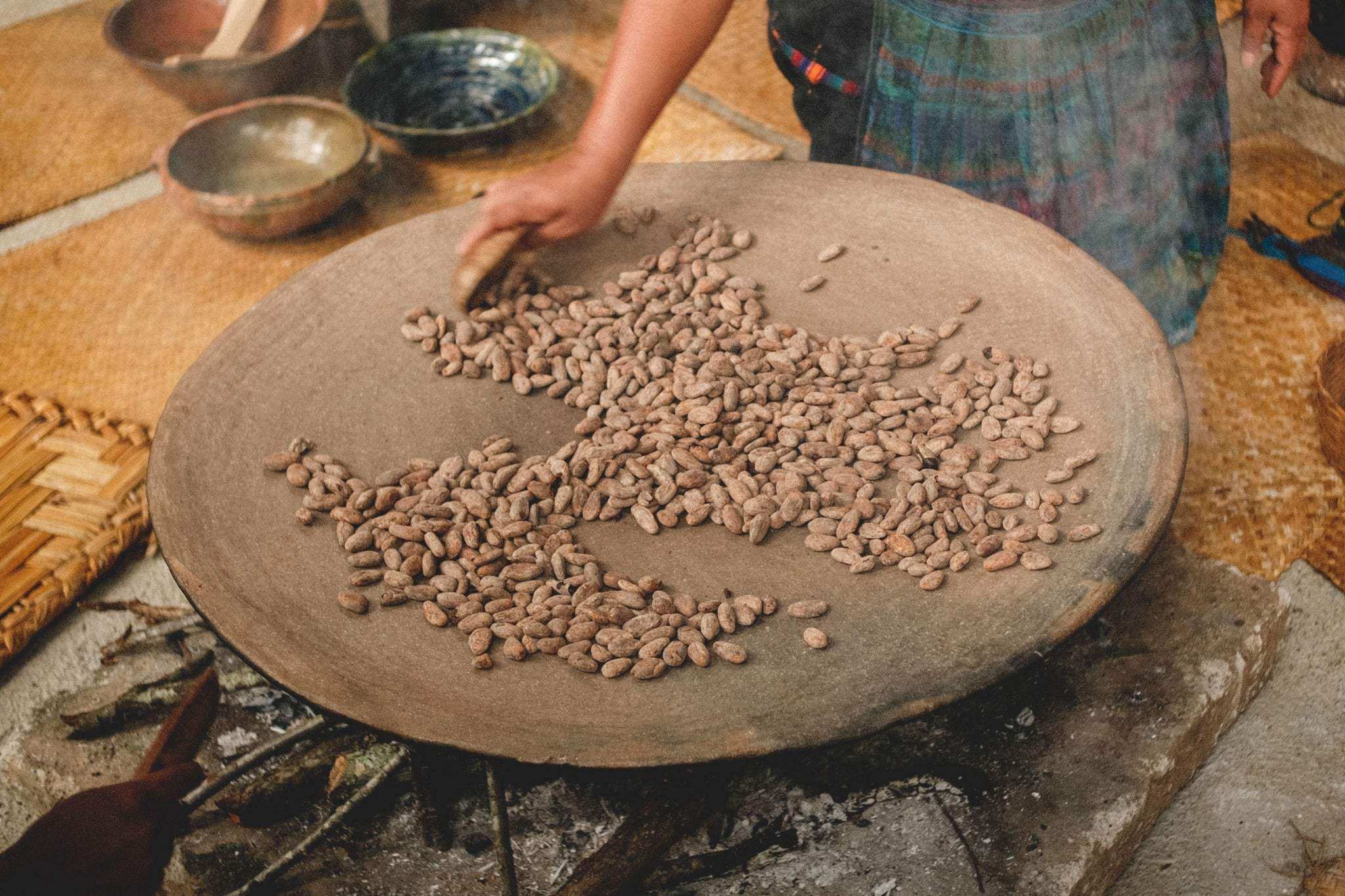 A person roasting pure cacao beans on a large round clay stove over an open fire, with woven mats and bowls visible in the background.