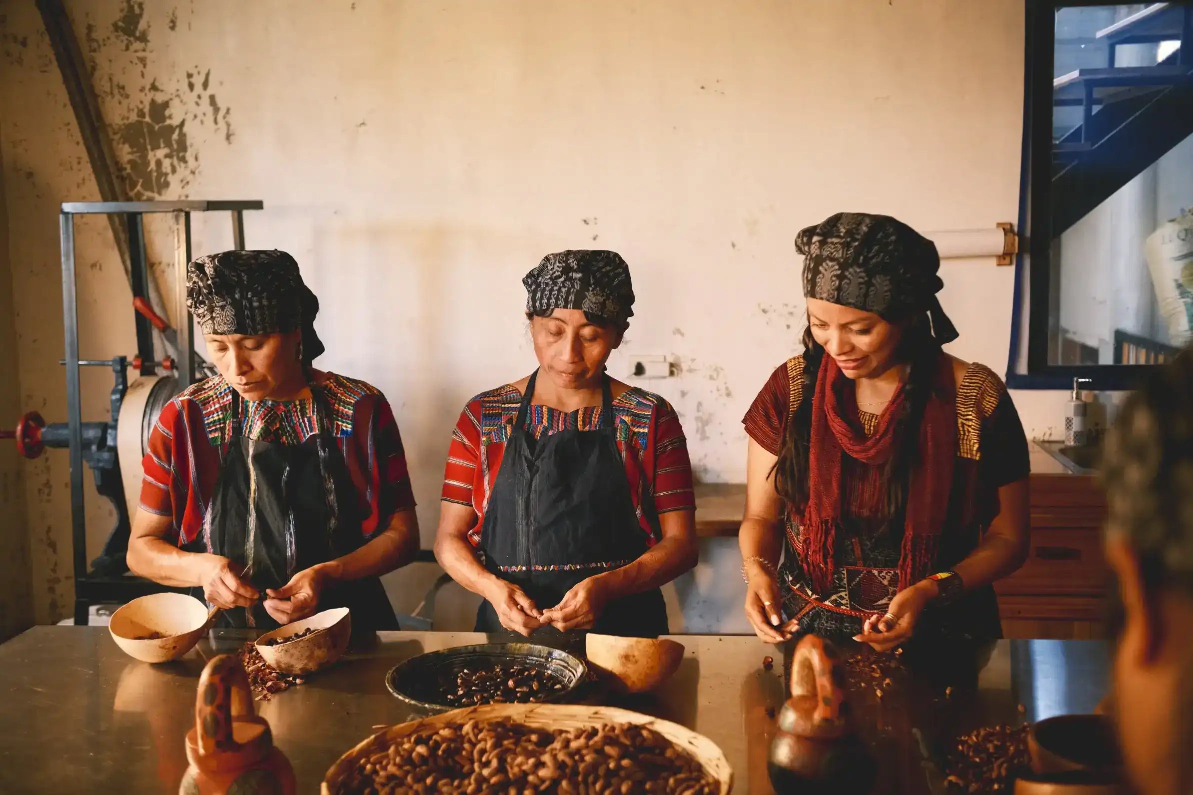 Three women in traditional Mayan clothing and headwear work at a table, handcrafting raw cacao beans in bowls and baskets to produce authentic ceremonial pure cacao powder inside a rustic indoor setting.